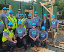 A group of construction workers in blue helmets pose together at a building site with wooden framing.