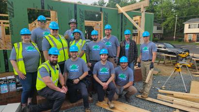 A group of construction workers in blue helmets pose together at a building site with wooden framing.