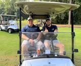 Two men in golf attire smile and sit in a golf cart on a sunny day, with another cart visible behind them.
