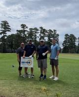 Four people standing on a golf course with a "Sponsored by DRY-PRO" sign, trees in the background.