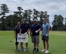 Four people standing on a golf course with a "Sponsored by DRY-PRO" sign, trees in the background.