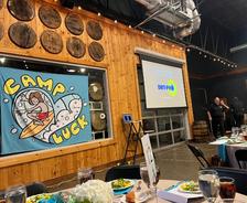 Indoor event with a "Camp Luck" banner, food on tables, and a screen displaying "DRYPRO.