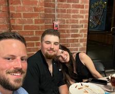 Three people smiling at a restaurant table with empty plates and glasses in front of a brick wall.