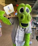 A green dragon mascot in a Charlotte Knights baseball jersey holds a sign while waving inside a stadium concourse.