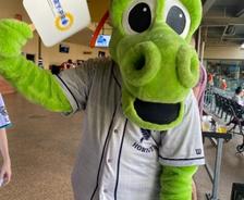 A green dragon mascot in a Charlotte Knights baseball jersey holds a sign while waving inside a stadium concourse.