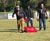 Three men play cornhole outside on grass, one tossing a beanbag, all wearing matching black shirts.