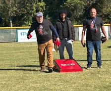 Three men play cornhole outside on grass, one tossing a beanbag, all wearing matching black shirts.