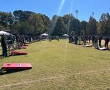 People playing cornhole on a grassy field in sunlight, with red boards and tents in the background.