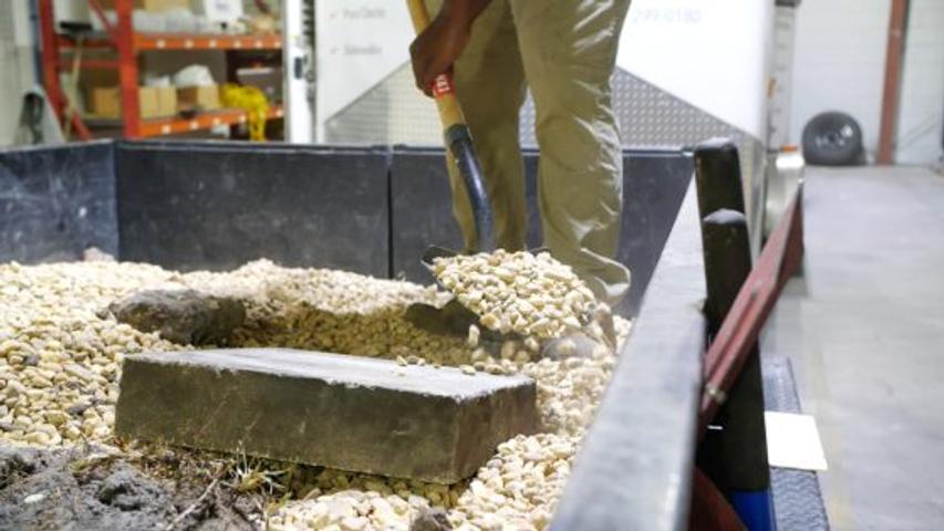 Person shoveling gravel from a trailer bed inside a warehouse with shelves in the background.