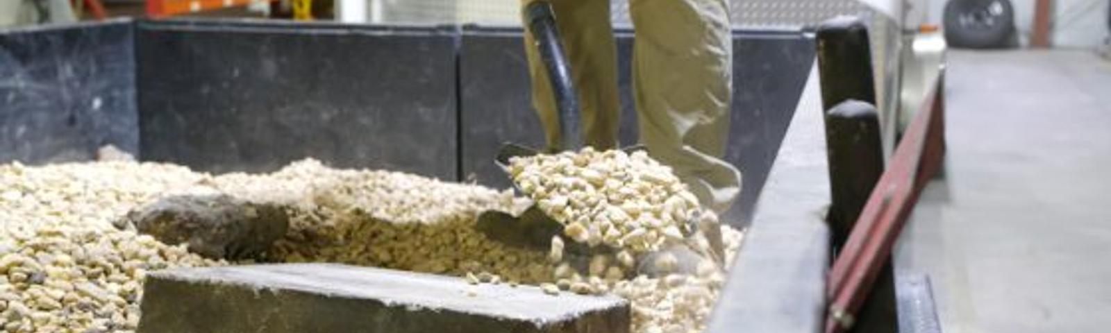 Person shoveling gravel from a trailer bed inside a warehouse with shelves in the background.