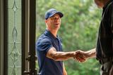 Smiling man in a blue polo and cap shakes hands with someone at a front door.