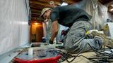 Two workers kneel on the floor installing equipment in a basement under construction with tools around them.
