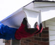 Person wearing red gloves installing a white rain gutter on a house with brick siding.