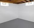 White basement corner with gray floor and exposed wooden ceiling beams, featuring two small rectangular windows.
