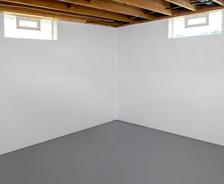 White basement corner with gray floor and exposed wooden ceiling beams, featuring two small rectangular windows.