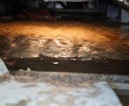 Wet, muddy ground inside a dimly lit crawl space shows clear signs of moisture, with water reflecting the light.