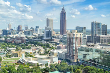 Skyline of a modern city with tall buildings, greenery, and a partly cloudy blue sky.