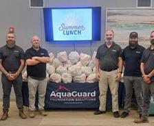 Group of six men standing by a table with "AquaGuard Foundation Solutions" banner and bags, under a "Summer Lunch" sign.