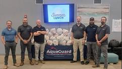 Group of six men standing by a table with "AquaGuard Foundation Solutions" banner and bags, under a "Summer Lunch" sign.