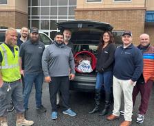 A group of people posing in front of an open car trunk filled with items, standing outside a building.