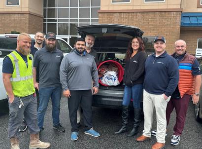 A group of people posing in front of an open car trunk filled with items, standing outside a building.