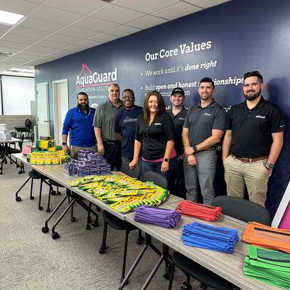 Group of people standing behind tables with school supplies at AquaGuard Foundation Solutions office.