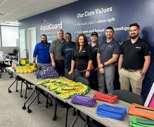 Six people standing by tables filled with school supplies in an office setting.