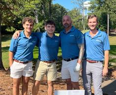 Four men in blue shirts pose together on a golf course, with trees in the background.