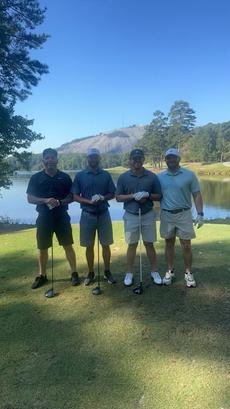 Four men with golf clubs stand on a course in front of a lake and wooded hill on a sunny day.