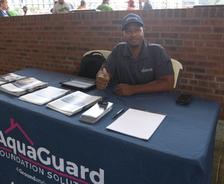 A man sits at a table with "AquaGuard Foundation Solutions" banner, giving a thumbs up.