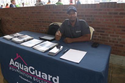 A man sits at a table with "AquaGuard Foundation Solutions" banner, giving a thumbs up.