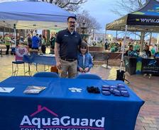 Man and child at AquaGuard Foundation Solutions booth at an outdoor event, surrounded by other stalls and people.