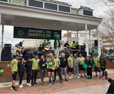 A group of people in green shirts pose in front of a stage with musicians at a "Shamrock Shuffle 5K" event.