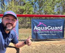 Man giving thumbs up near sign reading "AquaGuard Foundation Solutions, A Groundworks Company" on a chain-link fence.