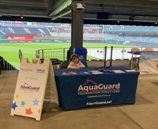 Child sits at an AquaGuard table with brochures at a stadium, next to a Make-A-Wish event sign.