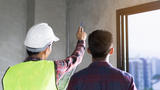 A construction worker in a hard hat points while showing plans to another person near a window.