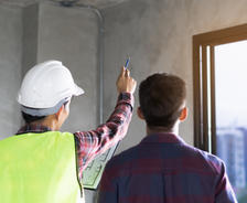 A construction worker in a hard hat points while showing plans to another person near a window.
