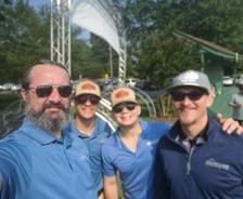 Four men in casual golf attire smiling outdoors, standing together for a group selfie on a sunny day.