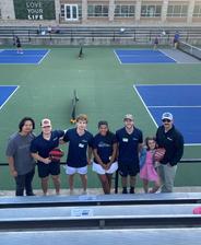 AquaGuard team members and family posing with pickleball paddles on a court, bleachers and others playing in the background.