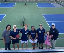 AquaGuard team members and family posing with pickleball paddles on a court, bleachers and others playing in the background.