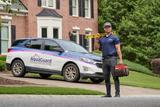 A worker holding a level and tool bag stands in front of a house and an AquaGuard Foundation Solutions car.