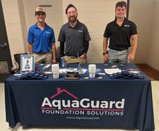 Three men stand behind an AquaGuard Foundation Solutions table display with promotional items and brochures.