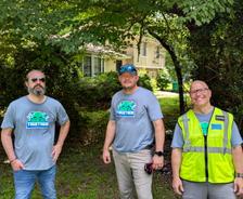 Three men in matching "Together" shirts stand outside near a house and trees on a sunny day.