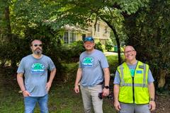 Three men in matching "Together" shirts stand outside near a house and trees on a sunny day.