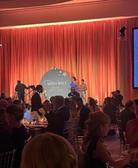 People seated at tables during a formal event with an orange curtain and "Wish Ball" sign on stage.