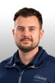 A man with short brown hair and a beard wearing a blue zip-up jacket and dark polo shirt, facing forward.