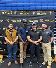 Four men stand smiling in front of a Carrollton Trojans backdrop in a gymnasium.