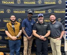 Four men stand smiling in front of a Carrollton Trojans backdrop in a gymnasium.