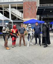 Five people pose outdoors, three in Star Wars costumes, two in casual clothes, by tents and a stadium building.