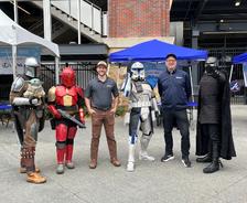 Five people pose outdoors, three in Star Wars costumes, two in casual clothes, by tents and a stadium building.